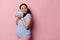 Happy gravid pregnant woman touches her face to clothes of future newborn baby, poses with eyes closed, isolated on pink