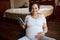 Happy gravid woman, keeping her hand on her pregnant tummy, smiling at camera, relaxing with a smartphone on a yoga mat