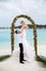 Happy groom holds bride on his hands under wedding ceremony arch on tropical island beach on Maldives. Turquoise ocean