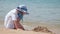 Happy hild girl in big hat and white dress playing alone with wet sand on sandy beach near clear sea lagoon water