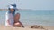 Happy hild girl in big hat and white dress playing alone with wet sand on sandy beach near clear sea lagoon water