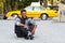 Happy hipster young man seated down on pavement road with crossed legs, isolated on a yellow blurred taxi background.