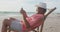 Happy hispanic senior man relaxing on sunbed on beach at sunset, using smartphone