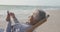 Happy hispanic senior woman relaxing on sunbed on beach at sunset, using smartphone