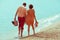 Happy honeymoon vacation concept. Young happy married couple of hipsters walking on beach holding each other and holding their