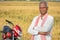 Happy Indian farmer with arms crossed standing with bike infront of the agriculture farmland - Concept of good crop yield and