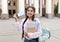 Happy indian student removing medical mask, posing outdoors with notepads near university building