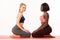 Happy international girls in sportswear sitting against each other on the red yoga mat, isolated over white background