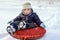 Happy joyful beautiful child boy rides from the mountain on red tubing in winter
