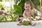 Happy and joyful infant baby at tropical vacation. Eats and drinks green young coconut. Sits on a ground in the jungles