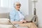 a happy, joyful old lady is sitting on a beige sofa with glasses on her face and headphones on her head working at home