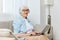 a happy, joyful old lady is sitting on a beige sofa with glasses on her face and headphones on her head working at home