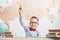 A happy, joyful schoolboy wears glasses, sits at a desk and raised his hand upwards