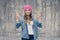 Happy joyful smiling hipster girl in casual clothes and pink hat standing against gray wall and showing victory sign, v-sign, gest