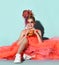 Happy kid girl with colorful dreadlocks in coral fatin dress, sneakers and sunglasses sitting on floor and eating cheeseburger