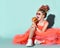 Happy kid girl with dreadlocks in coral fatin dress and sneakers is sitting on floor and eating cheeseburger looking aside