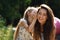 Happy kid girl whispering the secret to her laughing young mother in ear with fun face on summer green tree and grass background.