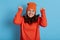 Happy lady dresses orange jumper and cap  keeps eyes closed and making hooray gesture  raising hands  feels very happy