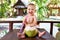 Happy and laughing infant baby at tropical vacation. Eats and drinks green young coconut. Sits on a wooden table