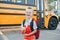 Happy laughing smiling Caucasian boy student by yellow bus on first September day. Hard of hearing child kid eating apple fruit at