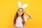 Happy little beautiful girl on the head with rabbit ears, shows a peaceful gesture on a yellow background