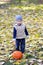 Happy little boy in cap and vest is standing with halloween pumpkin surrounded by fallen leaves.