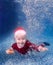 Happy little boy in Santa costume swimming underwater.