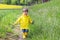 Happy little boy in yellow raincoat and muddy rubber boots running on dirt road through green grass near blooming rape seed field