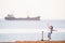 Happy little caucasian kid in helmet with scooter on sea beach with cargo ship on horizon