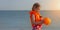 Happy little girl with blonde hair in kids swim vest with inflatable orange ball in hands, colorful seascape sunset