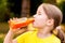 Happy little girl drinking healthy orange carrot juice from a glass bottle delighted, portrait, face closeup, outdoors. Healthy
