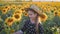 A happy little girl in the hat is smiling at the field of sunflowers at sunset time.