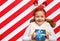 Happy little girl holding a Christmas present in her hands. Child in mittens headband on a red and white background