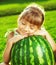 Happy little girl is hugging huge watermelon sitting on the green grass in summertime.