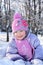 Happy little girl in pink scarf and hat lies in snow