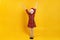 Happy little girl raised her hands up. Child in santa claus hat and red dress on a yellow background