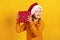 Happy little girl in santa hat holds christmas gift. Closeup portrait of a child on a yellow background