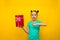 Happy little girl standing isolated over yellow background points at the red gift box in surprise. Looking camera