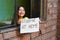 Happy little girl standing near the window and holding a sheet of paper that says `STUDYING AT HOME`.