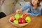 Happy little girl takes a pear from the bowl with variety fruits on table at home