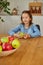 Happy little girl takes a pear from the bowl with variety fruits on table at home