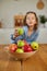 Happy little girl takes a pear from the bowl with variety fruits on table at home