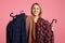 Happy lovely young woman holds clothes on hangers, rejoices new purchase, doesn`t know what to choose, going to spend money on new