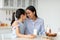 Happy loving asian mother and little daughter eating snacks in kitchen together, enjoying tasty cookies with milk