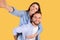 Happy loving european couple taking selfie together, cheerful young woman piggybacking her boyfriend, yellow background