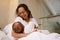 Happy loving mother smiles while connects with her baby, holds him in her arms, sitting on the bed in white bedchamber