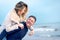 Happy man giving piggyback ride to his woman and laughing at beach. Smiling guy in love carrying on back her girlfriend