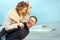 Happy man giving piggyback ride to his woman and laughing at beach. Smiling guy in love carrying on back her girlfriend