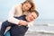 Happy man giving piggyback ride to his woman and laughing at beach. Smiling guy in love carrying on back her girlfriend