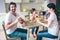 Happy man is sitting with his family at table. He holds cup of milk. Other members of family are eating food and talking